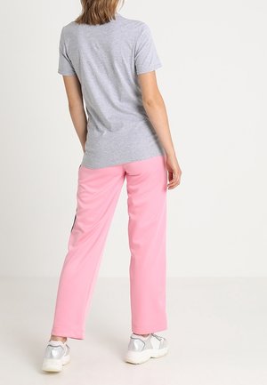 Person iført en lysegrå kortærmet t-shirt, lyserøde bukser og hvide sneakers, stående med ryggen til mod en ensfarvet baggrund.