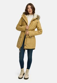 Brązowy płaszcz parka z sztucznym futerkiem, kieszeniami z przodu i talią ze sznurkiem; noszony na białym swetrze, w połączeniu z niebieskimi dżinsami i beżowymi botkami.