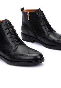 Des bottines en cuir noir présentent un laçage à l'avant, des détails de brogue, une fermeture éclair sur le côté et un petit talon empilé. Fini lisse avec un intérieur marron.