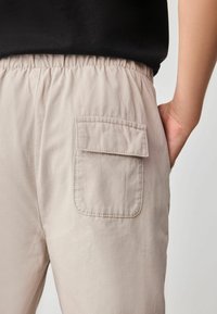 Helle beige Hose mit einem elastischen Bund und einer einzelnen aufgesetzten Tasche auf der rechten Rückseite, mit einer glatten, weichen Textur.
