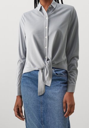 Femme portant une chemise boutonnée à rayures gris clair nouée à la taille, associée à une jupe en jean bleue, debout devant un fond uni.