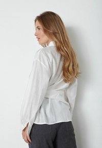 Biała, półprzezroczysta bluzka z długimi rękawami, zwężona w talii, z kołnierzykiem stojącym i subtelną teksturą. Połączona z szarymi spodniami.