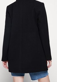 Cappotto in misto lana nero con una forma sartoriale, spacco singolo sul retro, maniche lunghe e superficie liscia, mostrato sopra pantaloni di jeans corti.