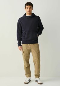 Tumesinine kapuutsiga dressipluus eesmise taskuga, kombineeritud beežide küljetaskutega ja elastsete mansettidega cargo-pükstega ning valgete tennistega.