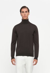 Man wearing a dark brown turtleneck sweater and white pants, standing against a plain white background.