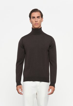 Mann iført en mørkebrun turtleneck-genser og hvite bukser, som står foran en ensfarget hvit bakgrunn.