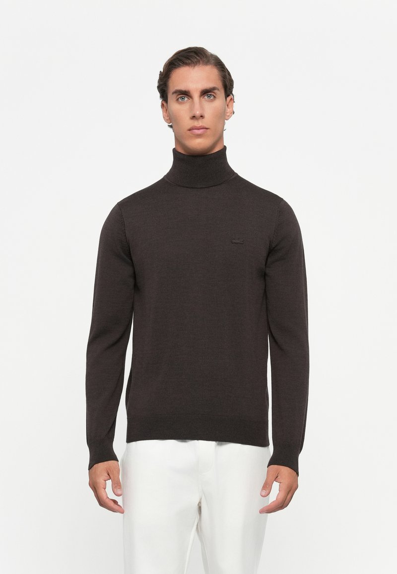 Man wearing a dark brown turtleneck sweater and white pants, standing against a plain white background.