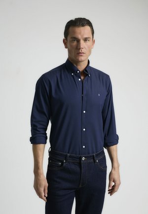 Camisa azul marino de botones con mangas arremangadas, confeccionada en una tela suave. Presenta un pequeño logotipo y botones blancos en contraste. Se lleva con jeans de mezclilla oscura.