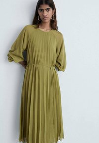 Rochie verde oliv pliată, cu decolteu rotund, mâneci lungi tip balon și talie evidențiată cu un braț. Fabricată dintr-un material ușor și fluid.