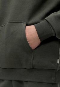 Χέρι που ακουμπάει στην μπροστινή τσέπη μιας σκούρας πράσινης φανέλας με ριμπ μανσέτες και τελείωμα.