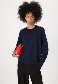 Mørkeblå hettegenser med preget mønster, rund halslinning og ribbestrikkede mansjetter, sammen med en liten rød lærveske.