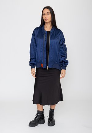 Frau steht, trägt eine dunkelblaue Bomberjacke, ein schwarzes Midikleid und schwarze Schnür-Kampfstiefel vor einem schlichten hellen Hintergrund.