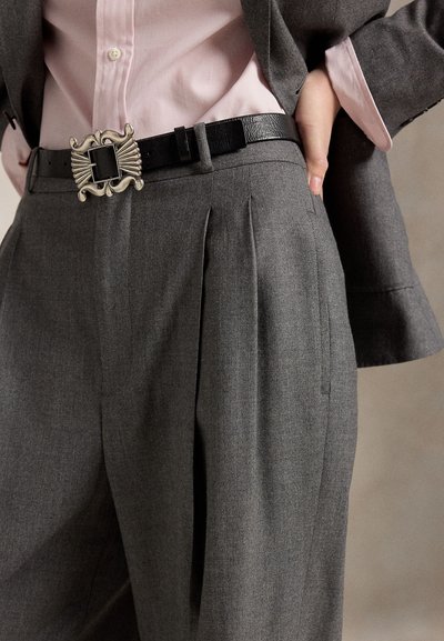 Maßgeschneiderte graue Hose mit Falten, getragen mit einem schwarzen Gürtel mit einer verzierten silbernen Schnalle, kombiniert mit einem pinken Hemd und einem grauen Blazer.