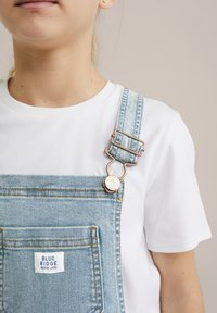 Svjetloplave traper kombinezone s prednjim džepom i mjedenim detaljima, uparenima s običnom bijelom majicom. Oznaka branda vidljiva na kombinezonima.