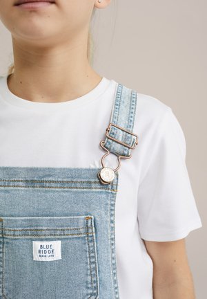 Salopete din denimul albastru deschis, cu un buzunar frontal și accesorii de culoare cupru, asociate cu un tricou alb simplu. Eticheta brandului este vizibilă pe salopetă.