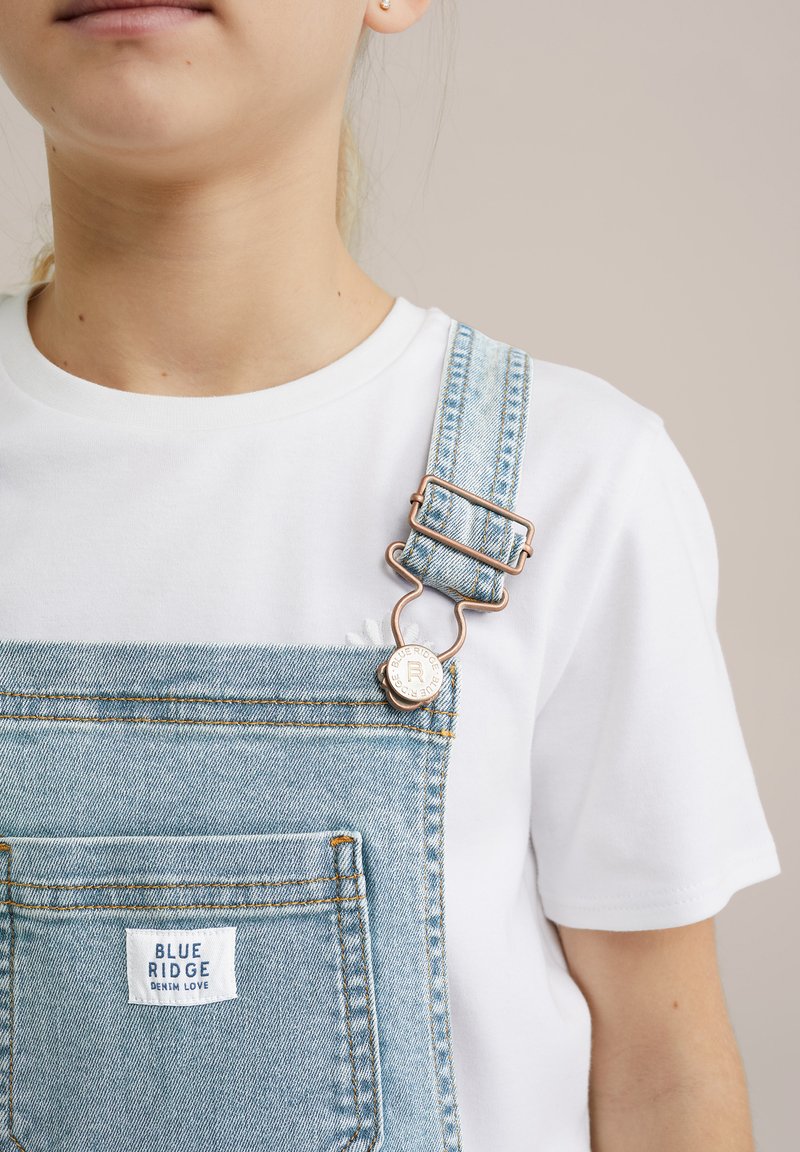 Svjetloplave traper kombinezone s prednjim džepom i mjedenim detaljima, uparenima s običnom bijelom majicom. Oznaka branda vidljiva na kombinezonima.