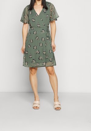 Woman wearing a knee-length green floral wrap dress with short flutter sleeves and beige open-toe slide sandals against a plain background.