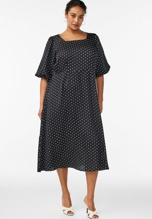 Kvinde iført en sort midikjole med hvide polkaprikker, pufærmer og firkantet udskæring, iført hvide åbne hæle.