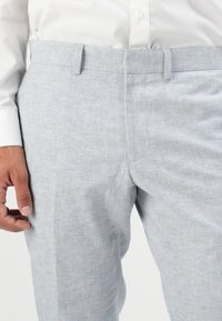 Χέρι άνδρα που ξεκουράζεται δίπλα σε ανοιχτό γκρι λινό παντελόνι συνδυασμένο με λευκό πουκάμισο μέσα στο παντελόνι.