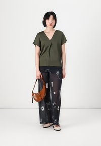 Blusa verde oliva a V con maniche corte, abbinata a pantaloni neri trasparenti con stampe floreali bianche. Borsa marrone a texture e scarpe basse beige.