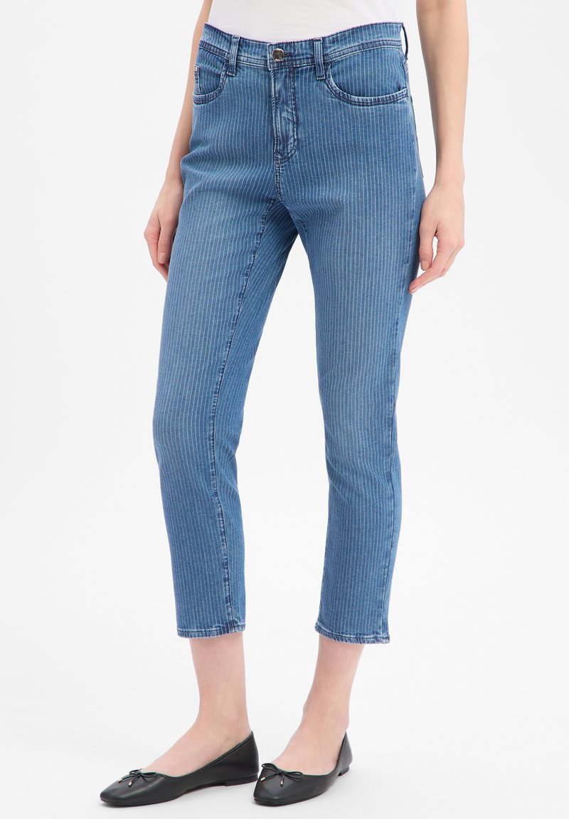 Kvinde iført blå, stribede kapri-jeans og sorte balletsko, stående med hænderne afslappet langs siden.