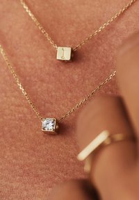 Gouden kettingen hebben kubistische hangers, waarvan er één is gegraveerd met een letter en de ander versierd is met een heldere edelsteen, op een gestructureerde huidachtergrond.