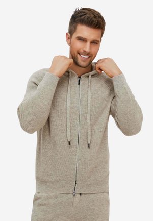 Ein lächelnder Mann richtet die Kapuze eines beigen, gestrickten Zip-up-Pullovers und trägt passende beigefarbene Hosen vor hellem Hintergrund.