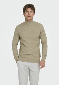 Beiger Pullover mit Reißverschlusskragen und gerippten Bündchen. Hergestellt aus weichem Material, mit feiner Textur und tailliertem Schnitt.