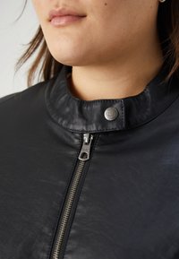 Svart läderjacka med hög krage, utrustad med silverdetaljer, sidodragkedja och en texturerad yta. Anmärkningsvärd knappar vid kragen.