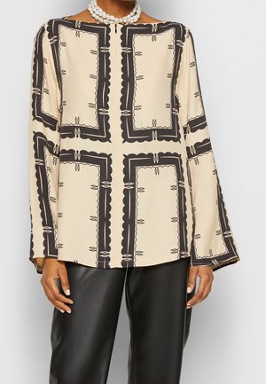 Vrouw draagt een beige blouse met lange mouwen met zwarte rechthoekige patronen en zwarte leren broek, geaccentueerd met een parelketting.