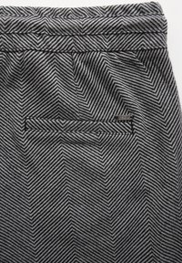 Poche arrière d'un pantalon gris et noir à motif chevrons avec une petite étiquette de marque métallique cousue près du bord de la poche.