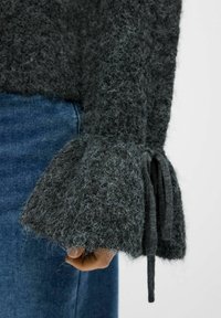 Mørkegrå strikket sweater med fuzzy tekstur, der har klokkeærmer bundet med et matchende stof. Båret over blå denim.