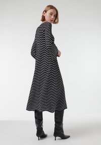 Vestido a rayas zigzag en blanco y negro con mangas largas, una silueta acampanada y un dobladillo que cae por debajo de la rodilla, combinado con botas negras hasta la rodilla.