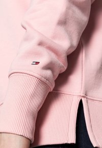 Světle růžová mikina s žebrovanými manžetami a lemem. Na rukávu má logo Tommy Hilfiger ve formě nášivky a po stranách na lemu jsou rozparky.