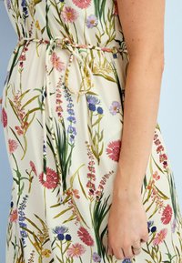 De jurk met bloemenprint heeft een crèmekleurige achtergrond met roze, blauwe en groene bloemen; is voorzien van een geknoopte tailleband en heeft een zachte textuur.