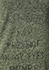 Camiseta verde oliva con un patrón texturizado que presenta texto en varias tipografías y adornos, incluidos acentos de tachuelas negras.
