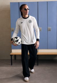 Fehér, hosszú ujjú, bordás póló fekete kiegészítőkkel, fekete, bő nadrág és fehér sportcipő. Fekete-fehér focilabdát tart.