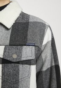 Personne portant une veste à carreaux noirs, gris et blancs avec un col en polaire blanc et une étiquette Superdry visible au-dessus de la poche poitrine.