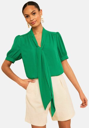 Žena s jasne zelenou blúzkou s dlhým predným viazaním a nafúknutými krátkymi rukávmi, v kombinácii s béžovými šortkami s vysokým pásom.