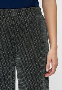 Svart bukser med et teksturert mønster i sølv, med elastisk midje. Stoffet ser glatt ut med en lett glans.