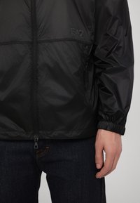 Veste zippée légère noire avec des poignets élastiques, arborant un logo de marque visible. Portée sur un jean en denim foncé, mettant en avant une texture lisse.