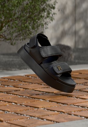 Μαύρο πλατφόρμα σανδάλι με δύο ρυθμιζόμενα λουριά και χρυσό λογότυπο, παρουσιάζεται σε εξωτερικό χώρο πάνω σε ξύλινη επιφάνεια με φυτεμένο φόντο.