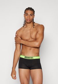 Black boxer briefs with a green Diesel logo waistband. Smooth fabric, fitted design, and mid-thigh length. Model shown with braided hair.