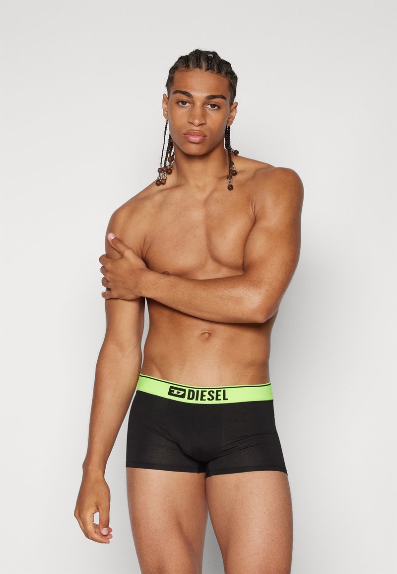 Black boxer briefs with a green Diesel logo waistband. Smooth fabric, fitted design, and mid-thigh length. Model shown with braided hair.