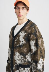 Pleteni kardigan u smešnim, belim i crnim kamuflažnim uzorku. Ima crni izrez u obliku slova V, prednje zatvaranje dugmadi i dva prednja džepa.