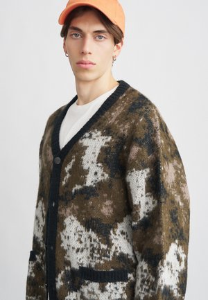 Cardigan lavorato a maglia in un pattern camouflage marrone, bianco e nero. Presenta un bordo a V nero, chiusura con bottoni sul davanti e due tasche frontali.