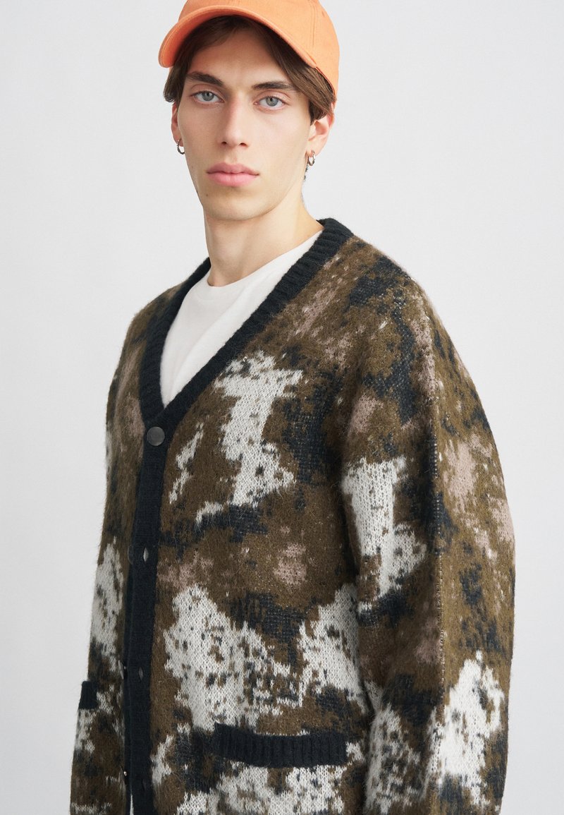 Pleteni kardigan u smešnim, belim i crnim kamuflažnim uzorku. Ima crni izrez u obliku slova V, prednje zatvaranje dugmadi i dva prednja džepa.