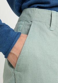 Pantalon vert clair avec une finition texturée et une poche latérale ; associé à un pull bleu, montrant une main dans la poche.