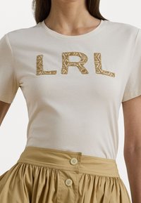 Trumpetų rankovėmis, kreminės spalvos marškinėliai su tekstūruotais auksiniais "LRL" rašmenimis; lygus audinys ir apvalus iškirpimas. Suderinta su mygtukais užsegama chaki spalvos sijonu.