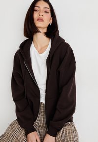 Brown zip-up hoodie with a spacious design, ribbed cuffs, and a front pocket, paired with a white t-shirt and plaid pants.